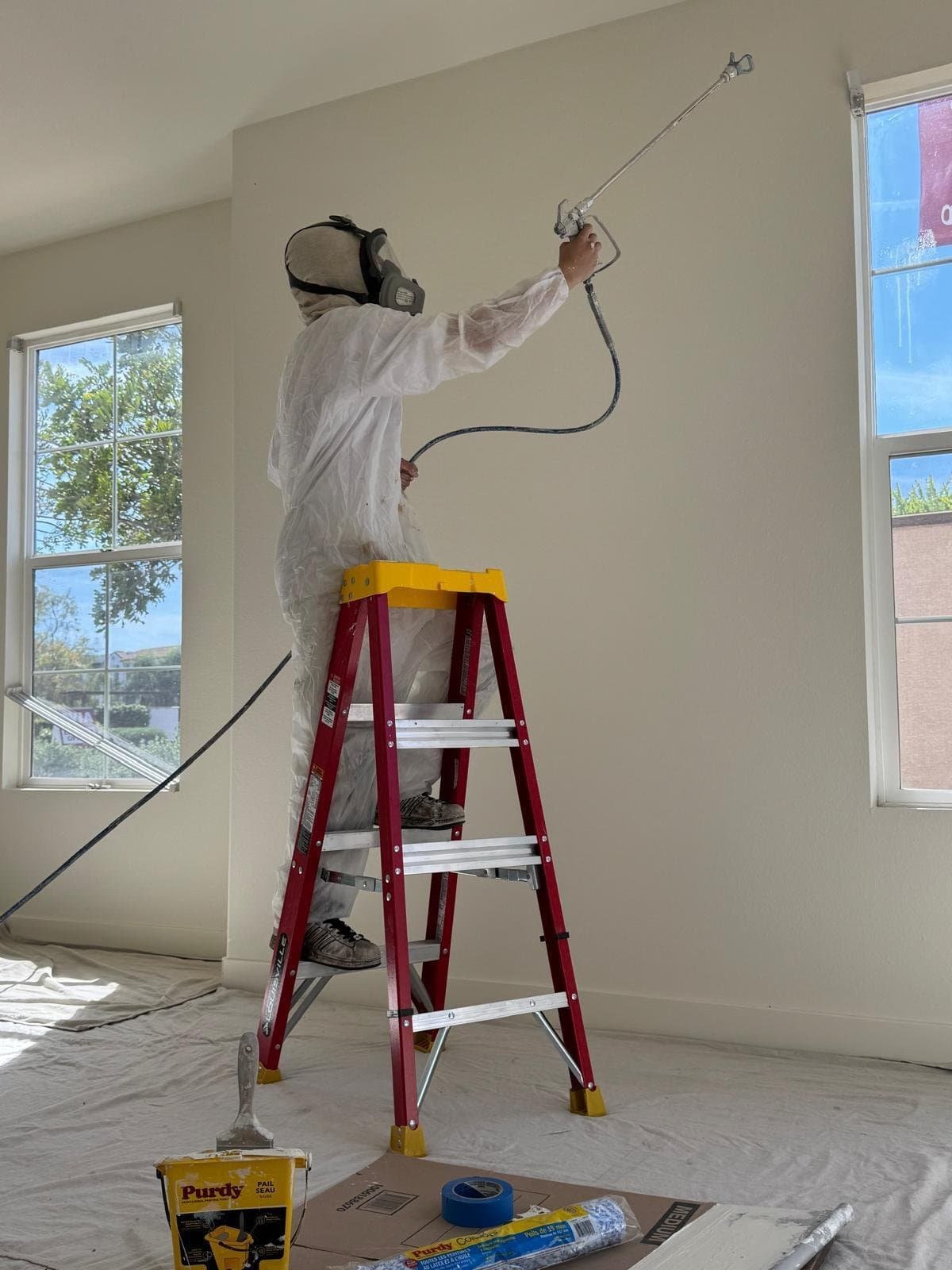 Worker painting interior wall with spray gun on ladder.